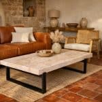 Idée déco salon tomette rouge : vue d’ensemble d’un salon chaleureux avec sol en tomettes rouge orangé, murs blanc cassé légèrement chauds, grand tapis berbère écru à motifs graphiques noir et écru, canapé en cuir cognac et fauteuil en rotin bois clair, table basse en travertin aux veines beige rosé et gris chaud, accents discrets en noir mat et laiton brossé, lumière douce créant une ambiance cosy et contemporaine, mettant en valeur la texture mate et la couleur du sol en terre cuite